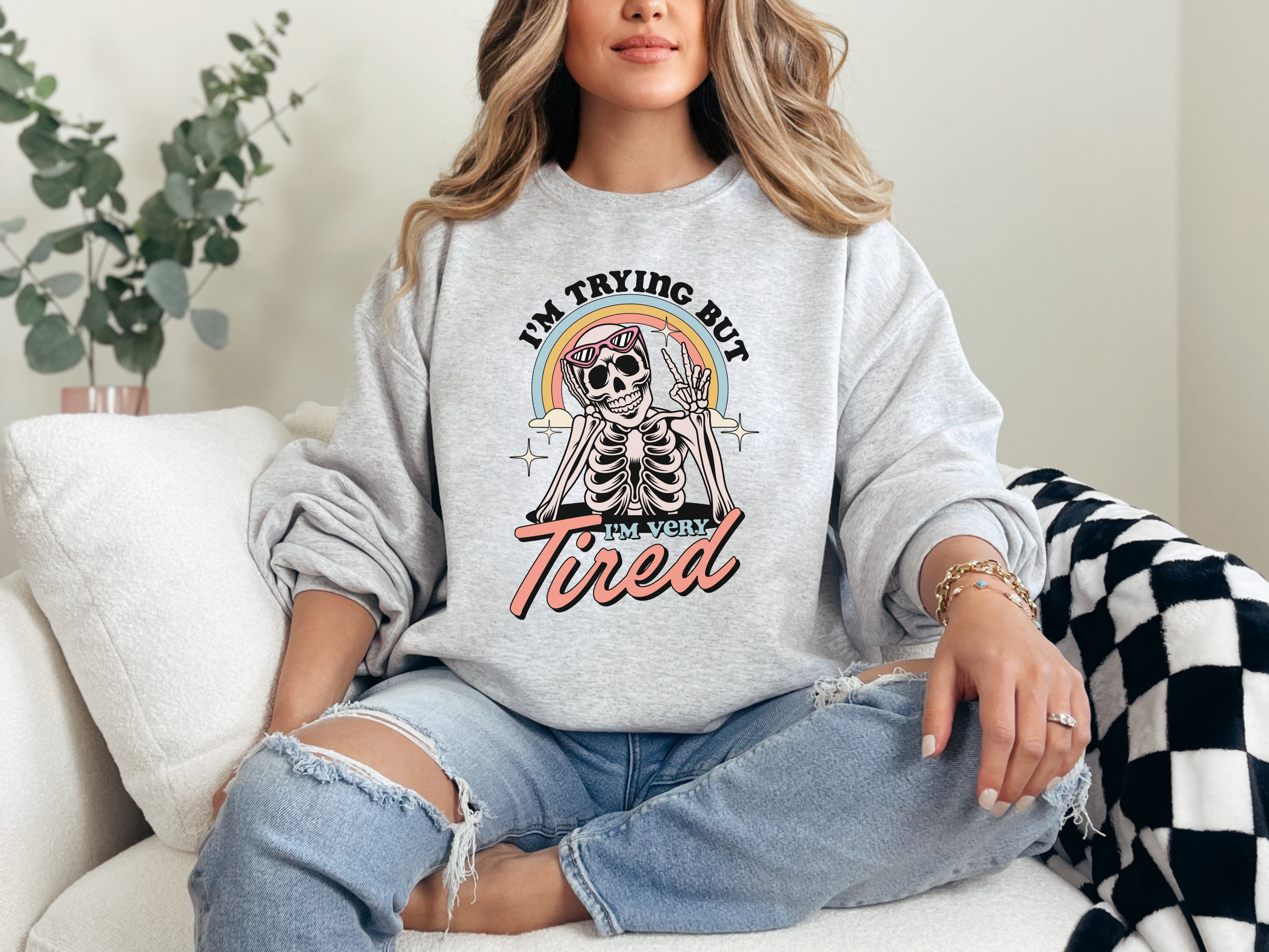 A sweatshirt with a design of a Skeleton wearing sun glasses in front of a rainbow giving a peace sign. The words "I'm trying but I'm very tired," written around it.