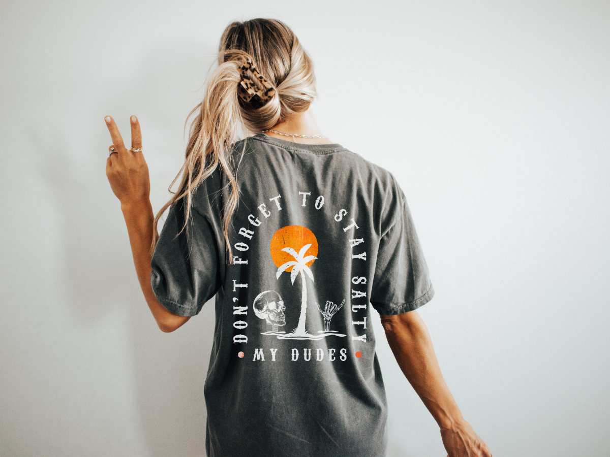 A dark grey t-shirt with the words don't forget to stay salty my dudes written in an arch surrounding an image of a skull and skeletal hand giving a hang loose sign around a palm tree at sunset.